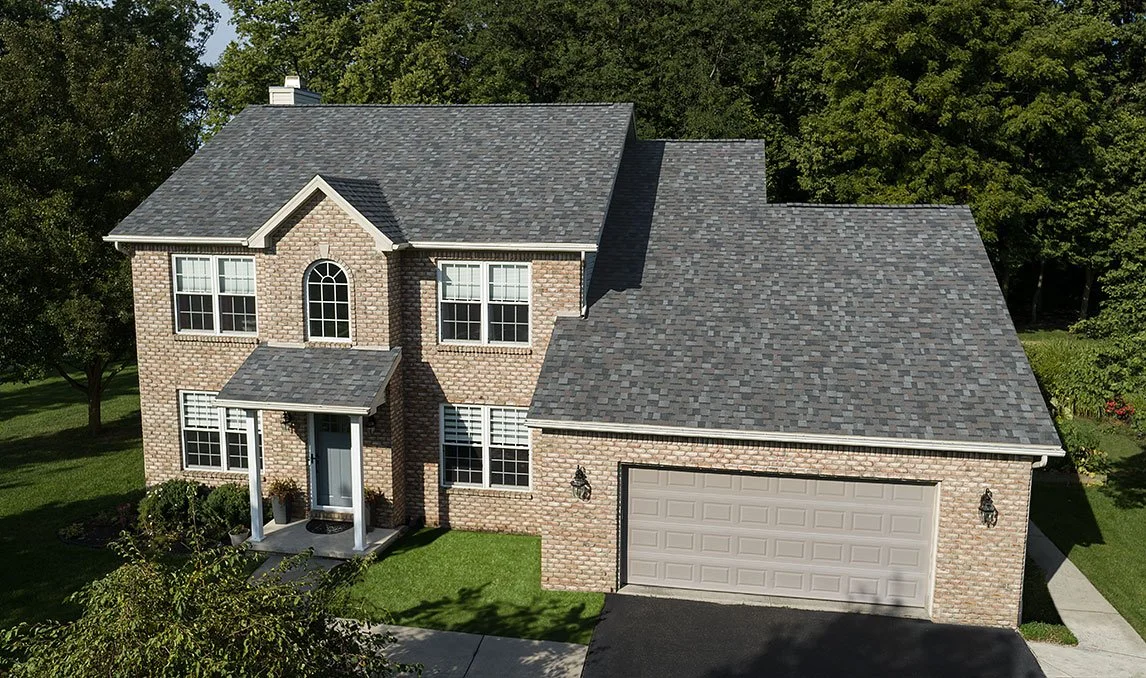 A two-story brick house with a gray shingle roof, a front porch with white columns, and a two-car garage. The house is surrounded by green grass and trees.