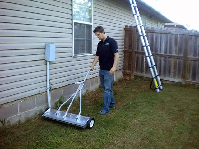 Man using a lawn roller to level the ground outside a house with beige siding and a wooden fence in the background.