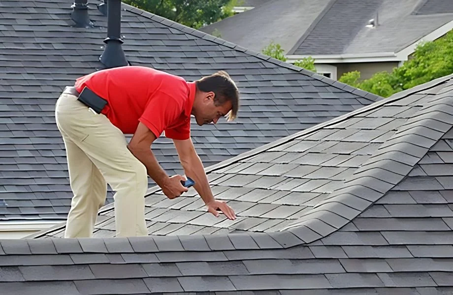 A man in a red shirt and beige pants inspecting a roof with gray shingles, using a tool in his right hand, on a sunny day with green trees in the background.