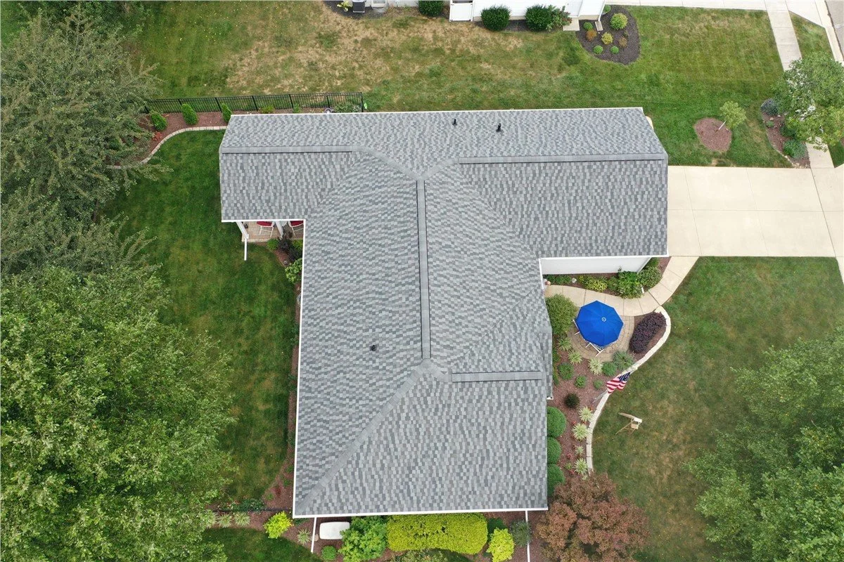 Aerial view of a house with a gray shingled roof, a well-maintained backyard with green grass, trees, a side patio with a blue umbrella, and a concrete driveway.
