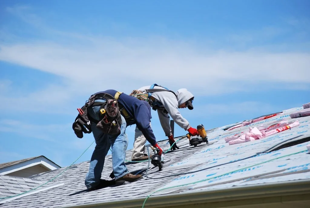 Two workers on a roof installing or repairing shingles, with tools and materials around them, under a partly cloudy sky.