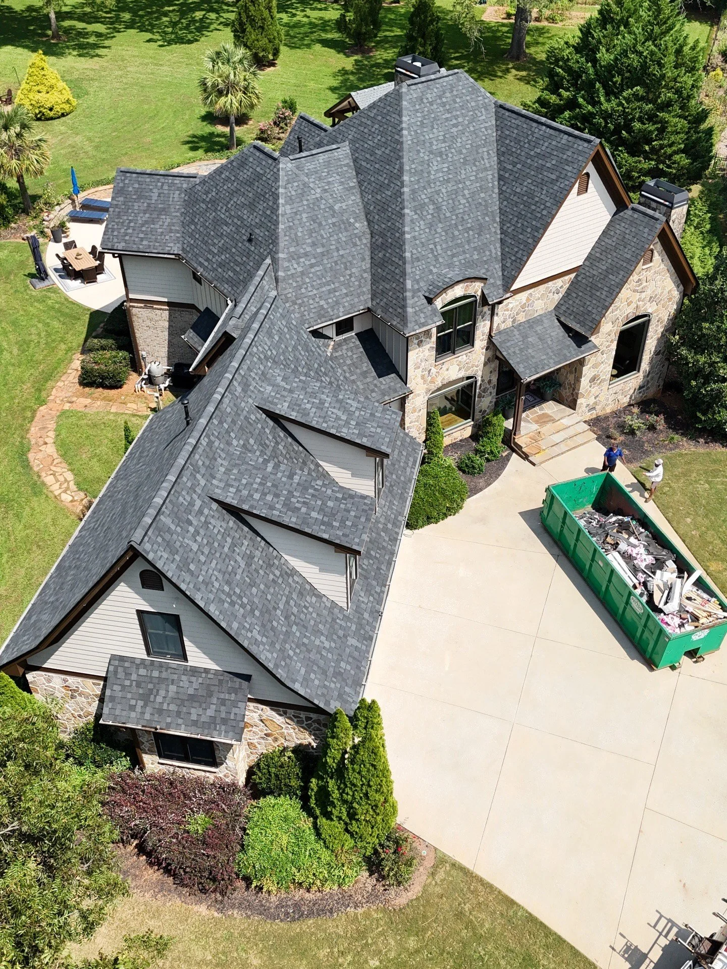 Aerial view of a house with gray shingle roof, stone and siding exterior, surrounded by a green lawn with trees, a patio with outdoor furniture, and a large green dumpster filled with debris.