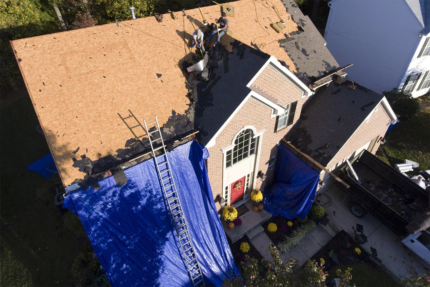 An aerial view of a house under construction, with the roof partially removed and workers installing new roofing materials, blue tarps covering parts of the house, and a ladder leaning against the roof.